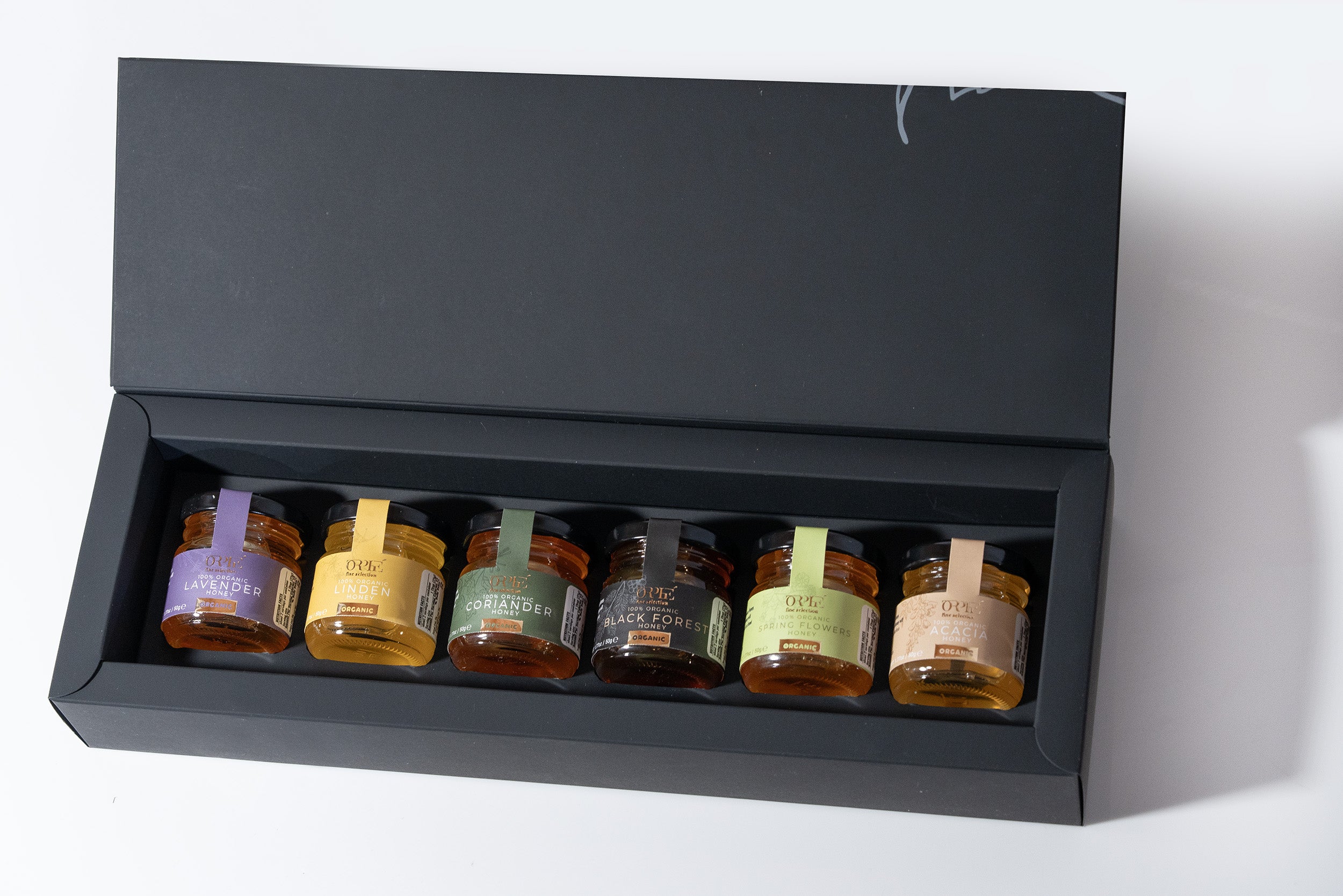 Set of six small jars with labels in a black display box on a white background, featuring lavender syrup and clover honey.