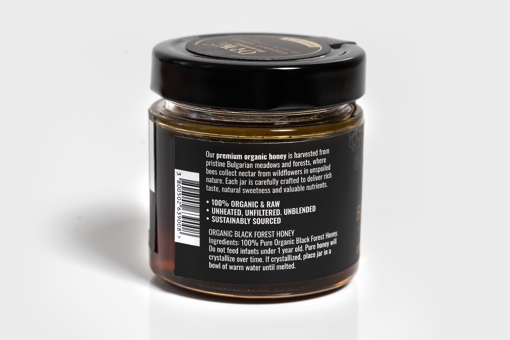 Clear glass jar filled with golden honey, topped with a sleek black lid. The jar is photographed against a clean white background, highlighting the rich color of the honey and the minimalist packaging.