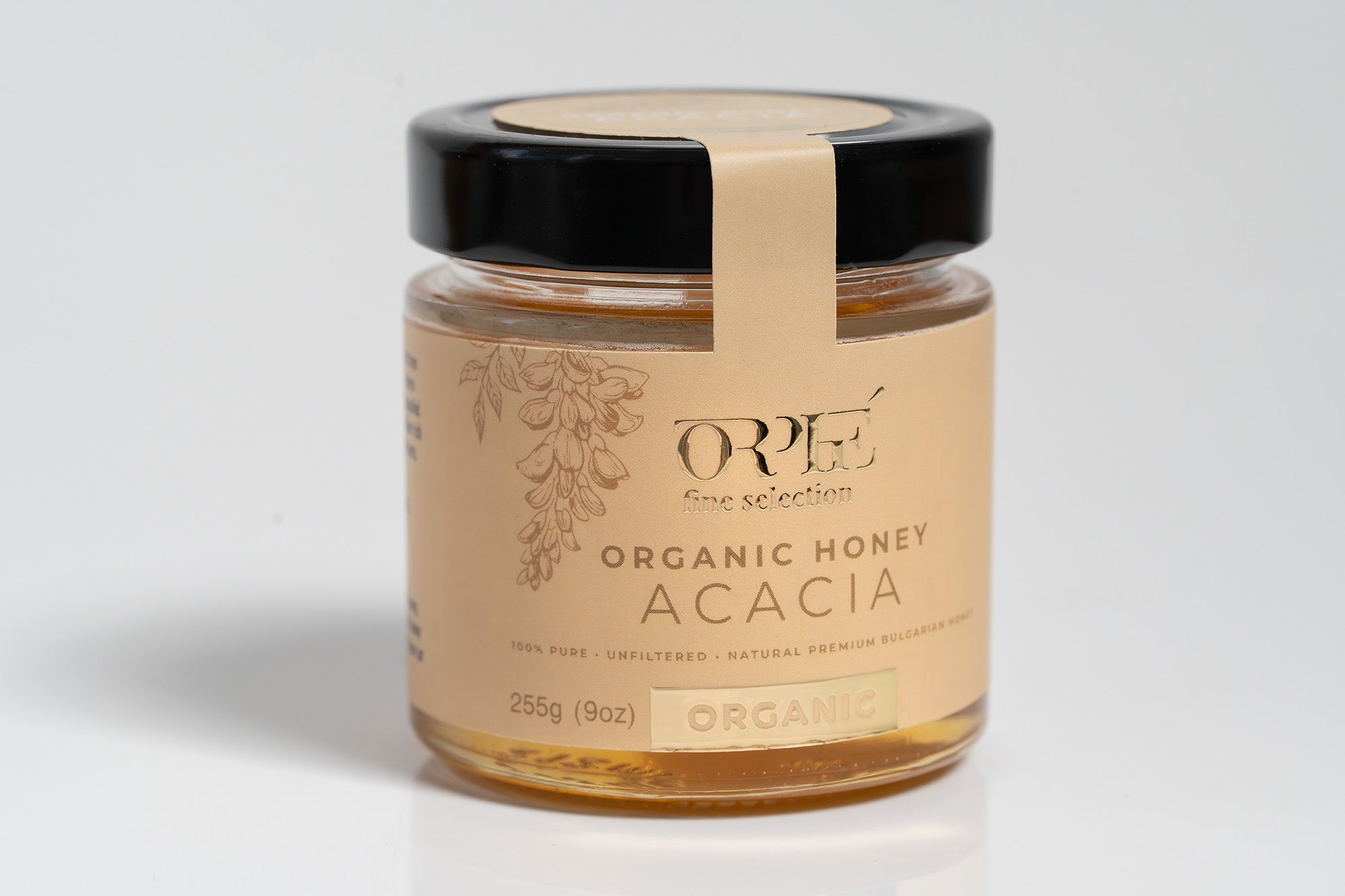 Clear glass jar of organic honey with a visible brand label featuring natural, earthy design elements. The jar is filled with golden amber honey and is set against a softly lit light background that enhances the product’s artisanal and wholesome appeal.