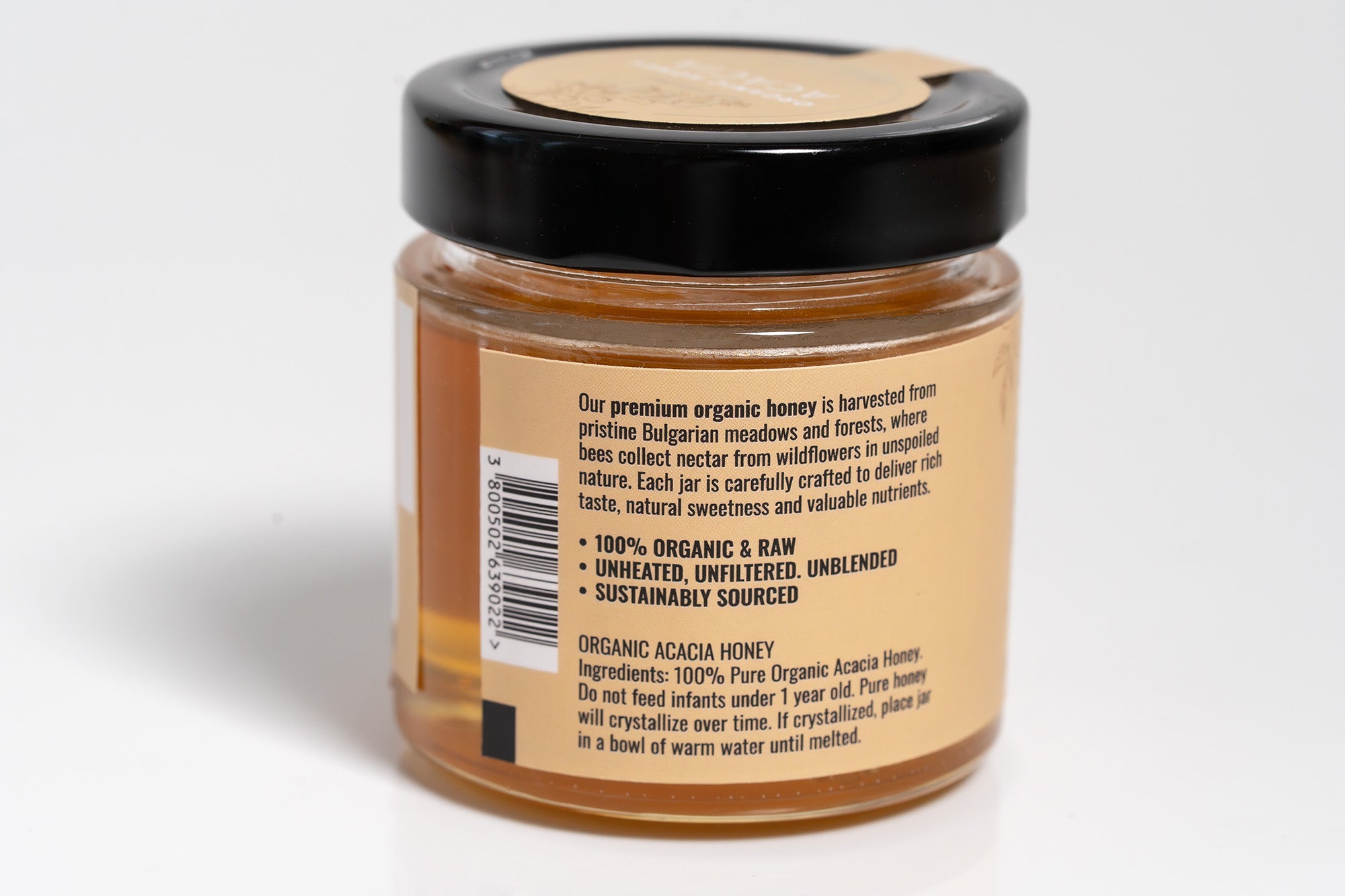 Glass jar filled with golden honey, featuring a clear and colorful label with product information. The jar is photographed against a bright white background, emphasizing the rich color of the honey and the design of the label.