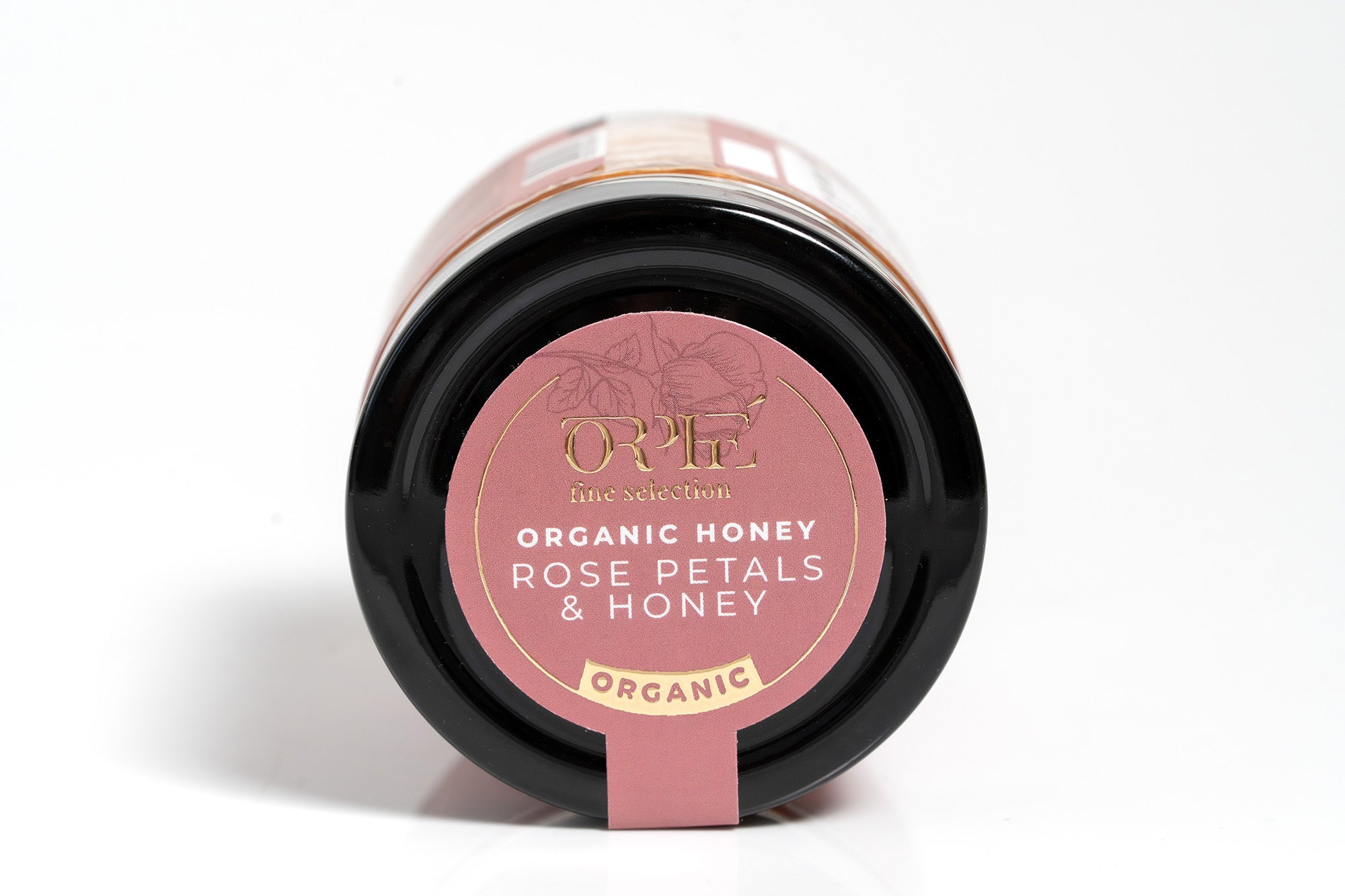 Clear glass jar of organic honey surrounded by fresh rose petals, showcasing the golden amber color of the honey inside. The jar is placed on a clean white background, emphasizing the natural and floral-inspired quality of the product.