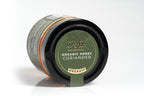 Clear glass jar of organic coriander-flavored honey with visible branding on the front label. The jar is filled with golden amber honey and is photographed against a clean white background, highlighting the product’s natural quality and distinctive flavor.