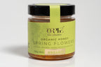 Glass jar of organic honey with a natural golden amber hue, featuring a green label with clean, eco-friendly design elements. The jar is sealed with a matching lid and is photographed against a bright white background, highlighting the purity and artisanal quality of the product.