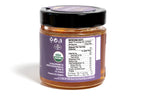 Clear glass jar of honey displaying the back side with a detailed nutrition label, showing product information and ingredients. The amber-colored honey is visible through the jar, and the product is photographed on a plain white background for a clean, professional look.